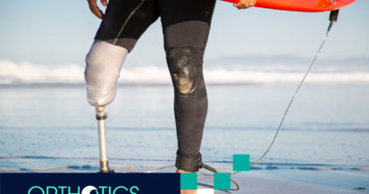 A male surfer with prosthetic legs is standing on beach sand holding a surf board