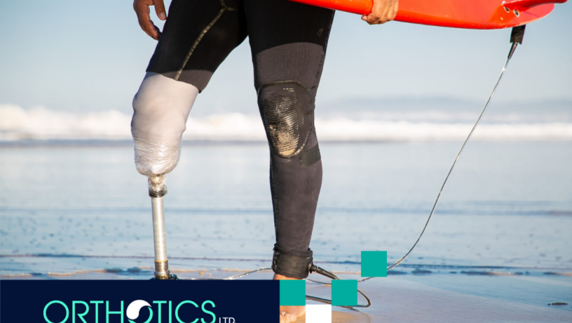 Caring for Your Prosthetic Limb - Featured A male surfer with prosthetic legs is standing on beach sand holding a surf board